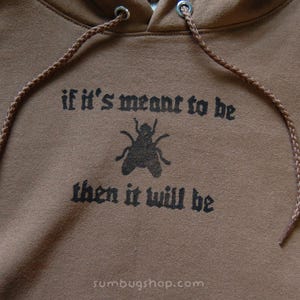 May include: Brown hoodie with a drawstring and a graphic of a fly and the text "if it's meant to be then it will be". The hoodie has a hood and a drawstring.