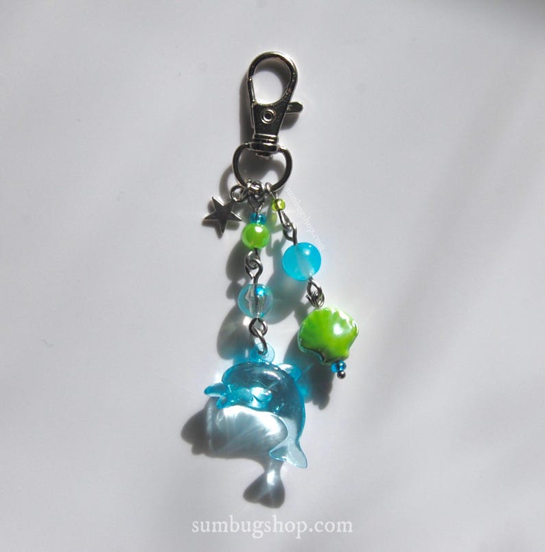 May include: A silver-tone keychain with a dolphin charm, star charm, and various blue and green beads. The dolphin is translucent blue. The keychain has a lobster clasp. The website sumbugshop.com is printed at the bottom.