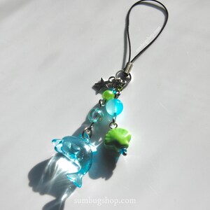 May include: A blue dolphin charm with a black cord, silver chain, and various beads. The charm includes a star, green and blue beads, and a green shell-shaped bead. The website sumbugshop.com is printed on the bottom.