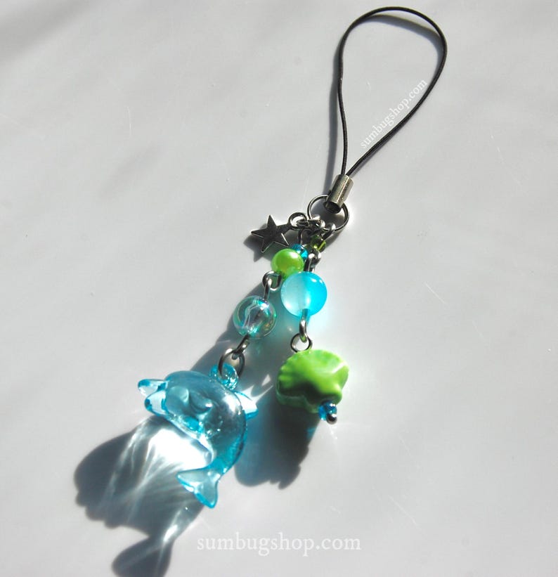 May include: A blue dolphin charm with a black cord, silver chain, and various beads. The charm includes a star, green and blue beads, and a green shell-shaped bead. The website sumbugshop.com is printed on the bottom.