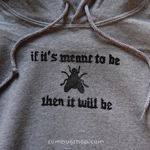 May include: A gray hooded sweatshirt with a drawstring. The front features the phrase "if it's meant to be then it will be" in black gothic lettering, with a black fly graphic. The website address sumbugshop.com is at the bottom.