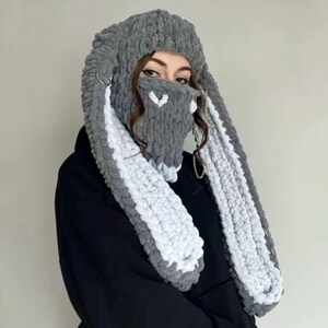 May include: Gray and white knitted hat with long bunny ear-shaped scarf. The hat has a face mask with a heart design.