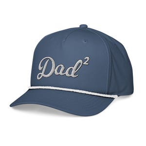 May include: A blue baseball cap with the word "Dad²" embroidered in white script. The cap has a white rope detail along the brim and a curved visor. The hat is a solid color and has a classic, casual style.