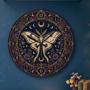 May include: A round wall clock featuring a central illustration of a cream-colored moth with intricate wing patterns, surrounded by a dark purple border with gold celestial motifs, floral accents, and black numerals from one to twelve.