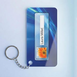 Puede incluir: Un llavero azul y blanco con una cadena y un anillo plateados. El llavero tiene un diseño rectangular con el nombre "Laura Miller" impreso verticalmente. Una pequeña imagen de una flor naranja está en la parte inferior.