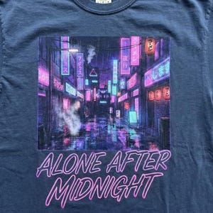 May include: Navy blue t-shirt featuring a neon-lit cityscape graphic with the words "ALONE AFTER MIDNIGHT" in a pink, stylized font. The graphic depicts a rainy street scene with Japanese lanterns and signs.