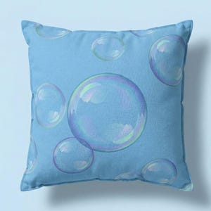 May include: Square cushion featuring a light blue backdrop adorned with a pattern of large, iridescent bubbles. The bubbles display a translucent, reflective quality, with hints of purple, green, and blue. The cushion is a soft, decorative item.