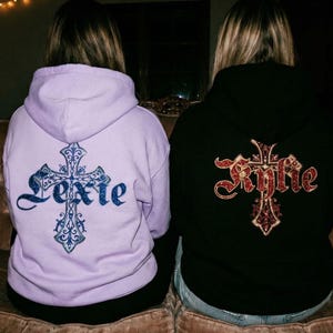 May include: Two people wearing hoodies with cross designs. One hoodie is lavender with a blue cross and the name "Lexie". The other is black with a red cross and the name "Kylie". They are sitting on a brown couch.