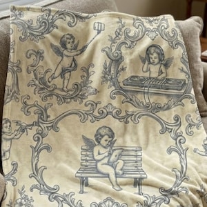 May include: A cream-colored throw blanket with a repeating pattern of cherubs in various poses, including using a tablet and taking selfies. The cherubs are in a gray-blue color, set against a backdrop of ornate scrollwork.