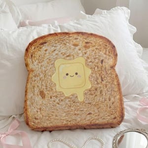 May include: A novelty cushion shaped like a slice of toast, with a smiling pat of butter. The pillow is brown with a yellow butter design. A pearl necklace, pink ribbons, and a hand mirror are also visible.