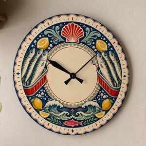 May include: A round wall clock featuring a cream-colored center and black hands. The border displays a decorative pattern with illustrations of sardines in red tins, yellow lemons, blue ocean waves, a red seashell, and a small red fish.