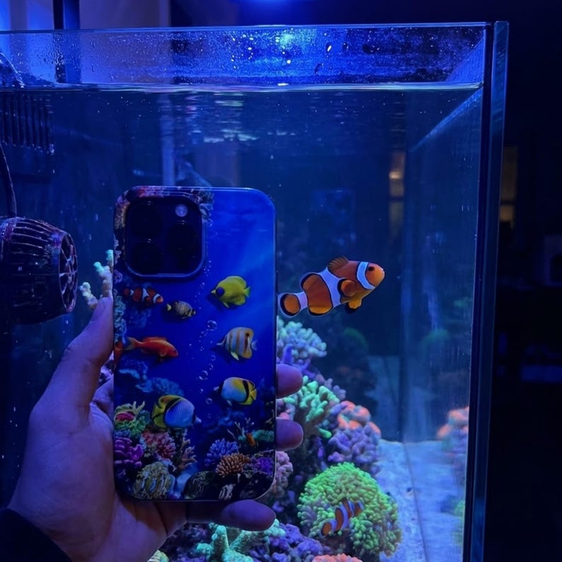 Pode incluir: Uma capa de telefone azul com uma vibrante cena subaqu&aacute;tica com peixes e corais coloridos. O design da capa inclui v&aacute;rias esp&eacute;cies de peixes, incluindo peixes-palha&ccedil;o, contra um fundo azul. A capa &eacute; segurada em frente a um aqu&aacute;rio.