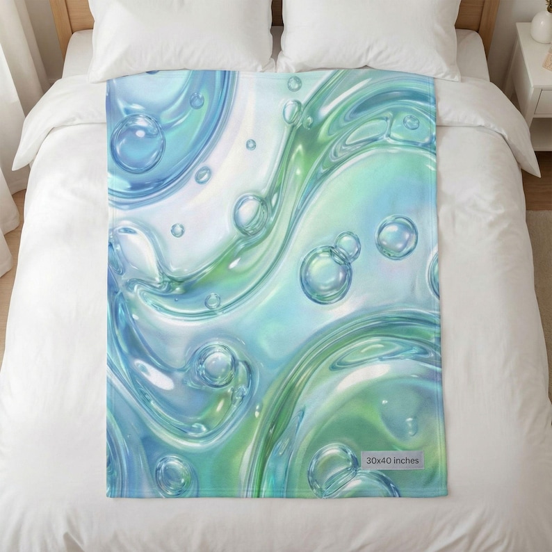 Może przedstawiać: Miękki koc z abstrakcyjnym wzorem wirującego niebieskiego i zielonego płynu z bąbelkami. Koc ma wymiary 76 x 102 cm. Wz&oacute;r przypomina wodę lub płynną, błyszczącą substancję.