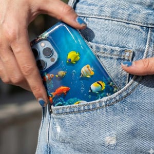 Pode incluir: Uma capa de telefone transparente com um design subaqu&aacute;tico vibrante, mostrando peixes coloridos, corais e bolhas. A capa &eacute; segurada em um bolso de ganga, destacando o tema aqu&aacute;tico. Os peixes incluem variedades amarelas, laranja e listradas.