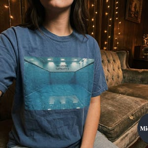 May include: A navy blue t-shirt featuring a graphic of a swimming pool with the text "NO DIVING CAPACITY 0". The shirt is a solid color and has a round neckline.
