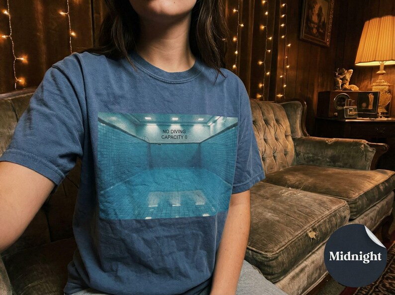 May include: A navy blue t-shirt featuring a graphic of a swimming pool with the text "NO DIVING CAPACITY 0". The shirt is a solid color and has a round neckline.