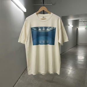 May include: Cream-colored t-shirt featuring a blue and white graphic of a pool with the text "NO DIVING CAPACITY". The shirt is hanging on a wooden hanger.