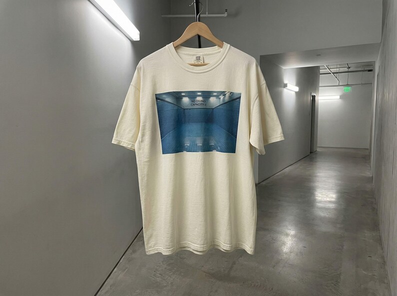 May include: Cream-colored t-shirt featuring a blue and white graphic of a pool with the text "NO DIVING CAPACITY". The shirt is hanging on a wooden hanger.