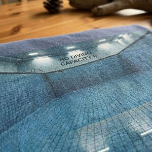 May include: A blue textile with a graphic of a pool interior, lit by overhead lights. The text "NO DIVING CAPACITY 0" is printed across the top. The image has a textured appearance.