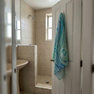 May include: A bathroom interior with beige tiled walls and a shower area. A white door on the right has a blue and green patterned towel hanging on it. A pedestal sink is visible on the left.
