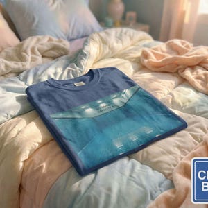 May include: A folded, China Blue t-shirt with a graphic design, resting on a bed with a patchwork quilt. The graphic on the shirt features a blue rectangular design with the text "NO DIVING CAPACITY 6". A China Blue color swatch is in the lower right corner.