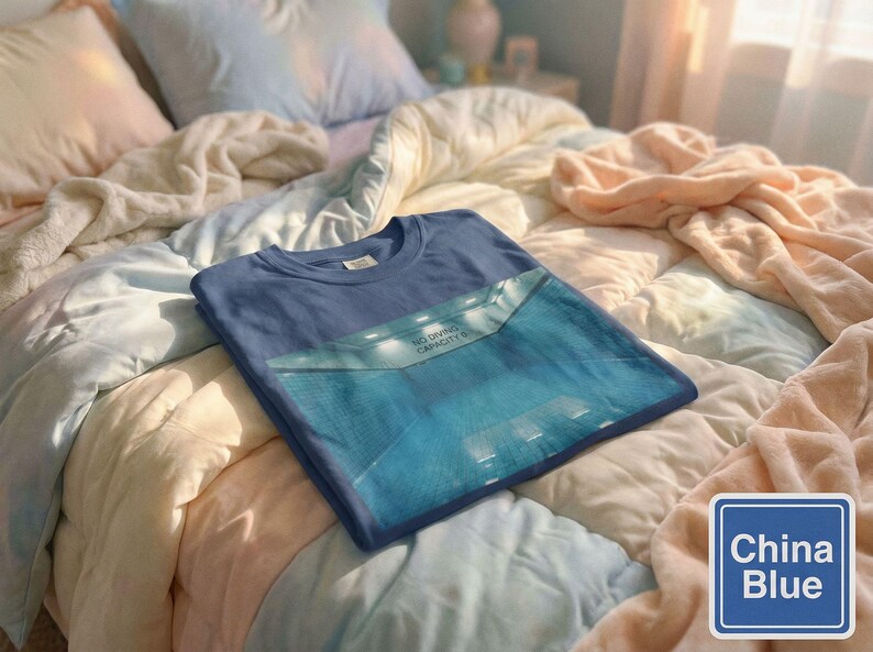 May include: A folded, China Blue t-shirt with a graphic design, resting on a bed with a patchwork quilt. The graphic on the shirt features a blue rectangular design with the text "NO DIVING CAPACITY 6". A China Blue color swatch is in the lower right corner.
