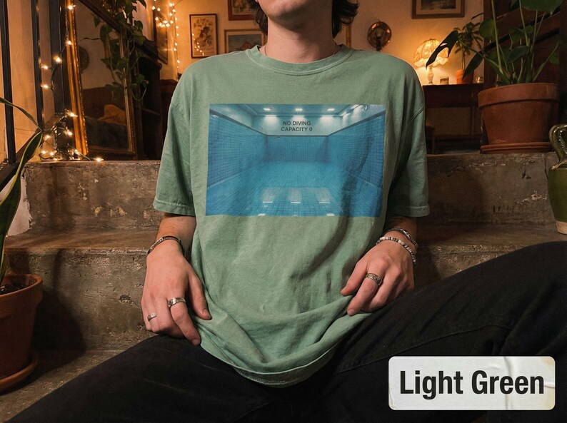 May include: Light green t-shirt with a rectangular graphic of a blue and white indoor pool scene. The text "NO DIVING CAPACITY 6" is above the pool. The shirt is a light green color.