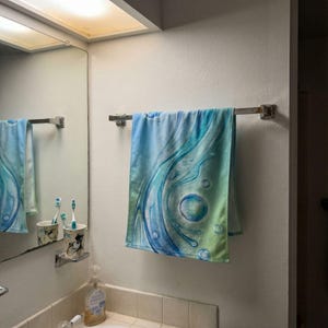 May include: A blue and green patterned towel hangs on a chrome towel rack in a bathroom. The towel features a flowing, abstract design with bubble-like shapes. The bathroom has a mirror, sink, and soap dispenser.