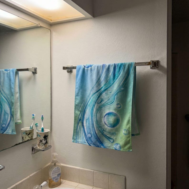 May include: A blue and green patterned towel hangs on a chrome towel rack in a bathroom. The towel features a flowing, abstract design with bubble-like shapes. The bathroom has a mirror, sink, and soap dispenser.