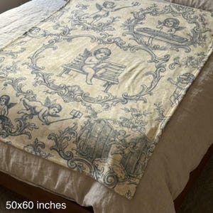 May include: A cream-colored throw blanket with a blue cherub and scroll design. The blanket measures 50 x 60 inches. The design features cherubs in various poses, including playing music and using a selfie stick, within ornate frames.