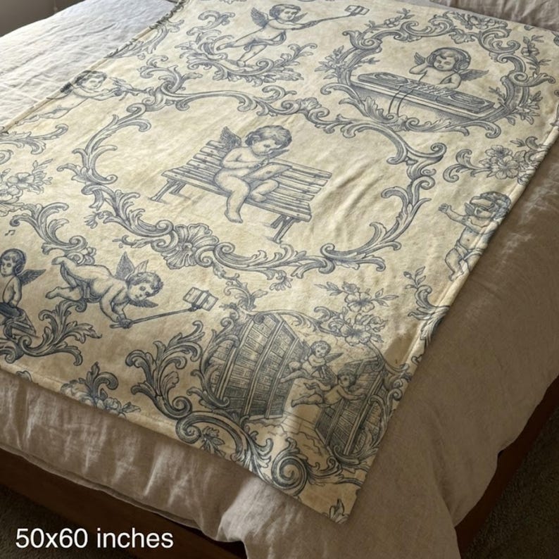 May include: A cream-colored throw blanket with a blue cherub and scroll design. The blanket measures 50 x 60 inches. The design features cherubs in various poses, including playing music and using a selfie stick, within ornate frames.