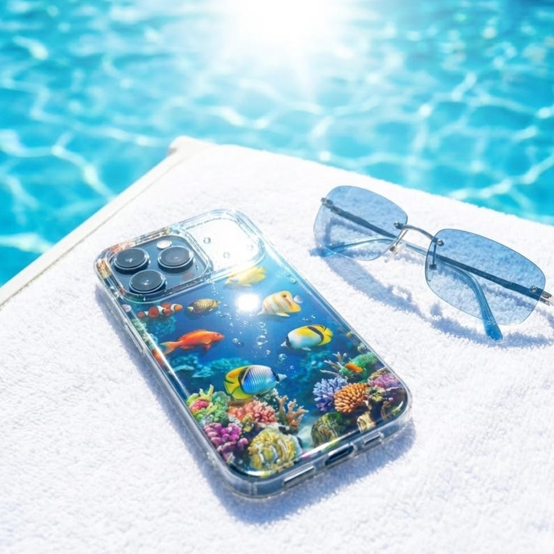 Pode incluir: Capa de telefone transparente com uma vibrante cena subaqu&aacute;tica com peixes e corais coloridos. A capa est&aacute; sobre uma toalha branca ao lado de &oacute;culos de sol azuis. Ao fundo, uma piscina.