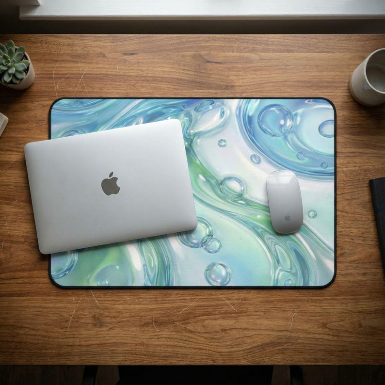 Puede incluir: Un ordenador port&aacute;til plateado y un rat&oacute;n blanco descansan sobre una alfombrilla rectangular con un dise&ntilde;o de burbujas azules y verdes. La alfombrilla est&aacute; sobre un escritorio de madera, con una peque&ntilde;a planta suculenta y una taza blanca.