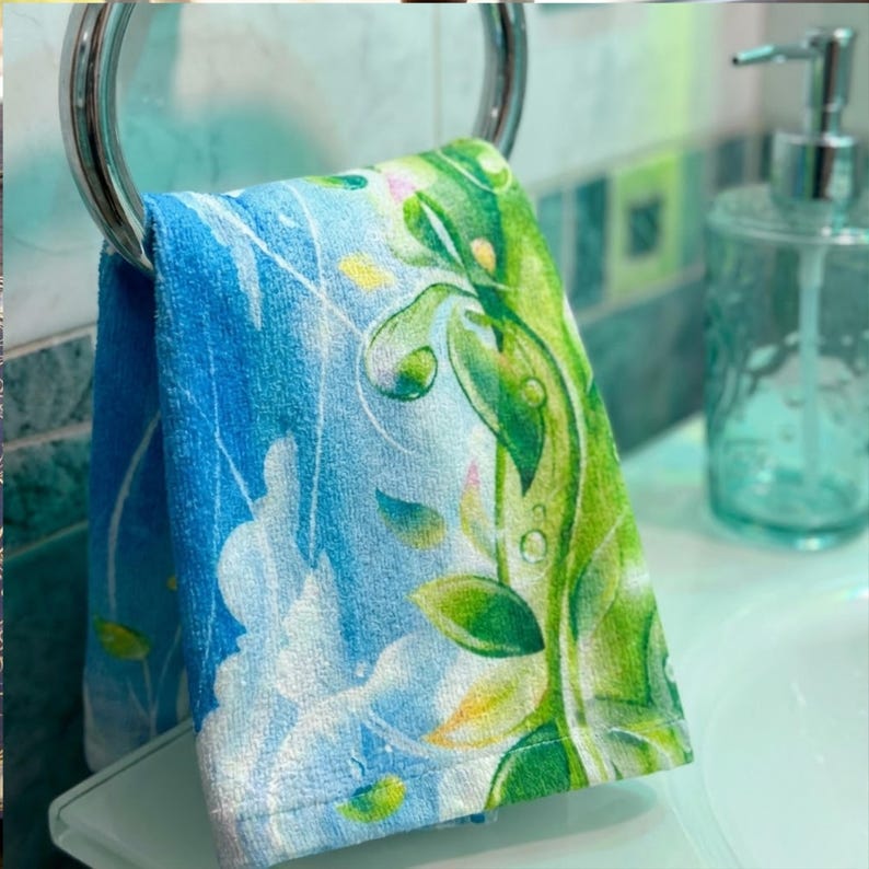 K&ouml;nnte beinhalten: Ein Handtuch mit blauem und gr&uuml;nem Blumenmuster h&auml;ngt an einem verchromten Handtuchring. Das Handtuch zeigt ein Design aus Bl&auml;ttern und Ranken in Gr&uuml;n und Gelb vor blauem Hintergrund. Im Hintergrund ist ein klarer Seifenspender zu sehen.