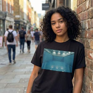 May include: Black t-shirt featuring a rectangular graphic of a blue indoor pool with the text "NO DIVING CAPACITY". The shirt is worn by a person with curly hair, standing in front of a brick wall.