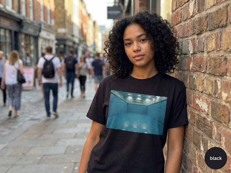 May include: Black t-shirt featuring a rectangular graphic of a blue indoor pool with the text "NO DIVING CAPACITY". The shirt is worn by a person with curly hair, standing in front of a brick wall.
