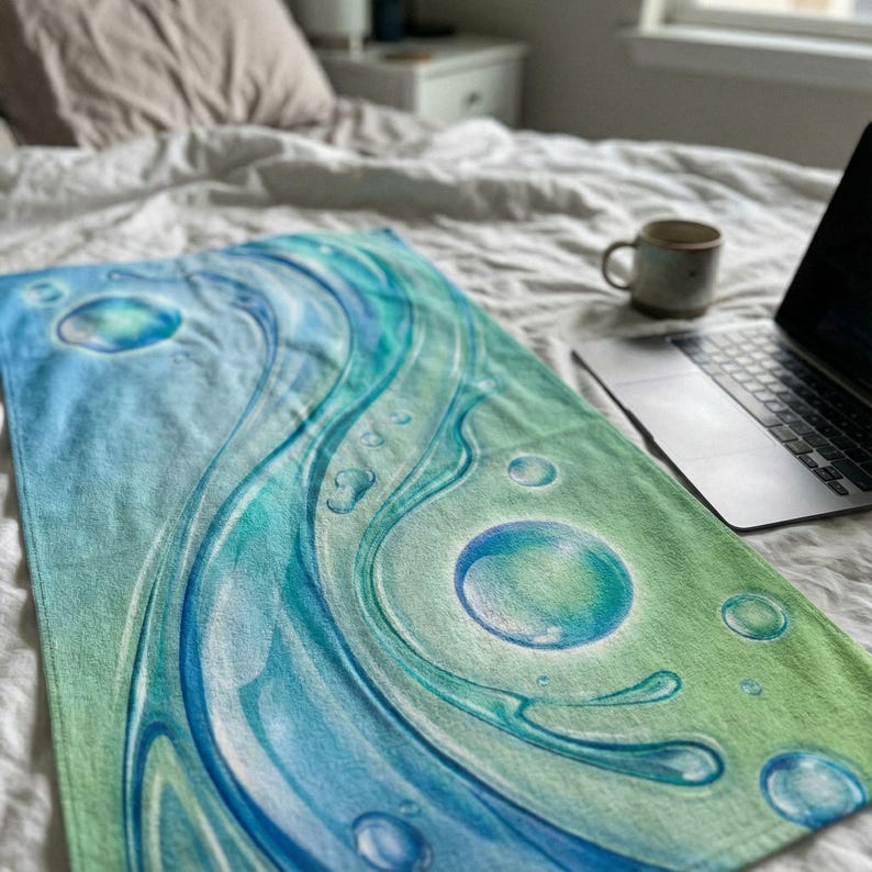 May include: A rectangular beach towel with a water-themed design. The towel features a gradient of blue and green hues with swirling water patterns and bubble accents. The towel is lying on a bed.