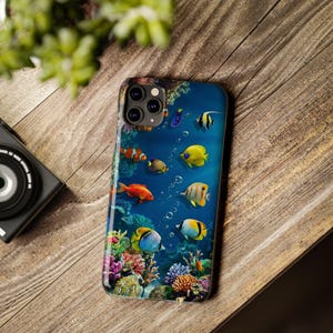 Pode incluir: Uma capa de smartphone com um design subaqu&aacute;tico vibrante, apresentando peixes coloridos, corais e bolhas. O design inclui peixes-palha&ccedil;o, peixes-cirurgi&atilde;o amarelos e outros peixes tropicais em um fundo azul. A capa est&aacute; em uma superf&iacute;cie de madeira.