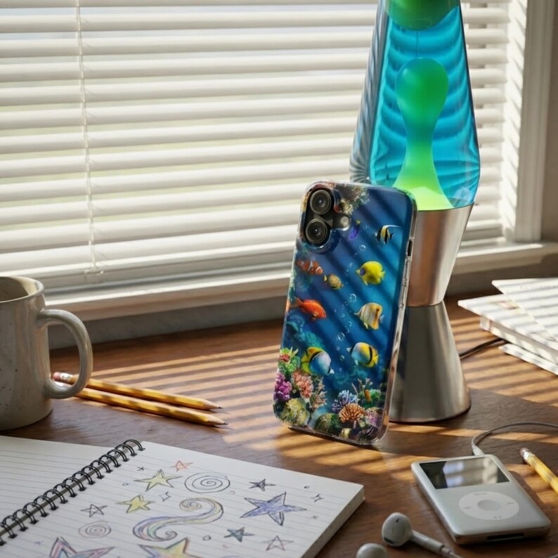 Pode incluir: Capa de telem&oacute;vel com um design subaqu&aacute;tico vibrante, apresentando peixes coloridos e corais contra um fundo azul. &Eacute; exibida perto de uma l&acirc;mpada de lava com l&iacute;quido azul e verde, um caderno com rabiscos de estrelas e uma caneca.