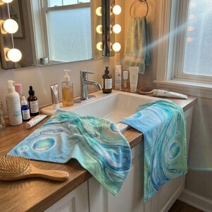 May include: A bathroom scene with a white sink, mirror, and wooden countertop. Two blue and green patterned towels are draped over the sink. Various skincare products and a wooden hairbrush are visible.