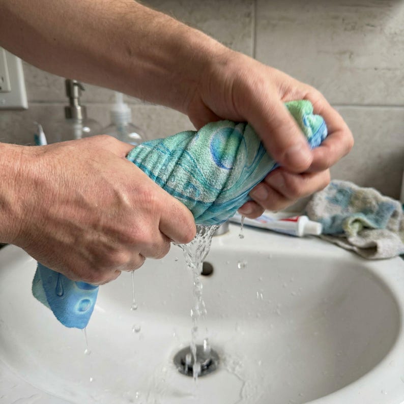 May include: A person squeezing a wet, patterned washcloth over a white sink. The washcloth is primarily blue and green, with water droplets falling from it. The sink is clean, and a partially visible tube of toothpaste is on the counter.