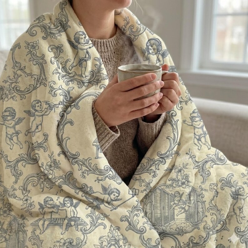 May include: A cream-colored blanket with a blue cherub and floral pattern. The blanket is draped over a person, who is holding a mug of a warm beverage. The blanket's design includes cherubs, floral elements, and decorative scrollwork.