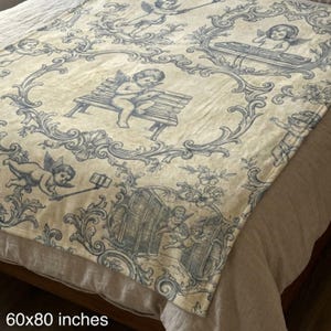 May include: A cream-colored throw blanket with a blue toile pattern featuring cherubs in various poses, including using a selfie stick and playing a record player. The blanket measures 60x80 inches.