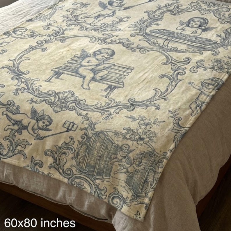 May include: A cream-colored throw blanket with a blue toile pattern featuring cherubs in various poses, including using a selfie stick and playing a record player. The blanket measures 60x80 inches.
