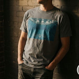 May include: A gray t-shirt with a blue and white graphic of a stadium. The shirt is short-sleeved and has a crew neck. The person is wearing blue jeans and is leaning against a brick wall.