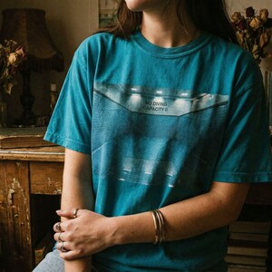 May include: Topaz blue t-shirt with a graphic design featuring the text "NO DIVING CAPACITY 0". The shirt is short-sleeved and has a relaxed fit. The image also shows a person wearing the shirt.