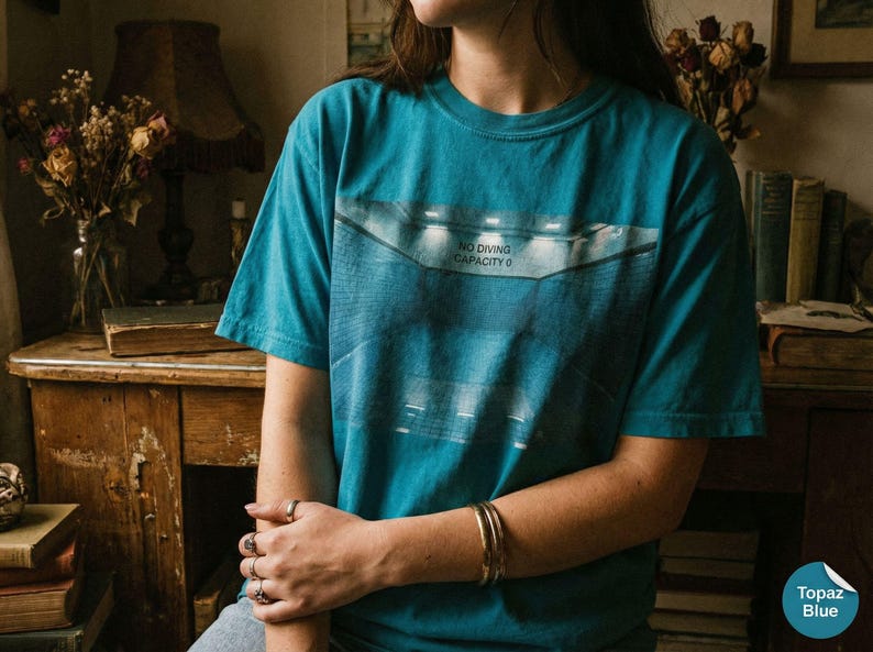 May include: Topaz blue t-shirt with a graphic design featuring the text "NO DIVING CAPACITY 0". The shirt is short-sleeved and has a relaxed fit. The image also shows a person wearing the shirt.