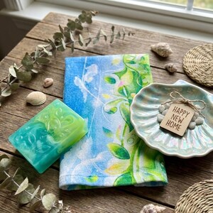 K&ouml;nnte beinhalten: Ein dekoratives Set mit einem blau-gr&uuml;n gemusterten Handtuch, einem Seifenst&uuml;ck und einer muschelf&ouml;rmigen Schale mit einem "Happy New Home"-Anh&auml;nger. Die Seife hat ein Wirbeldesign. Die Artikel sind auf einer Holzoberfl&auml;che angeordnet.