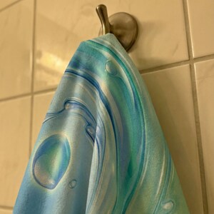 May include: A blue and teal patterned towel hangs from a silver hook on a tiled wall. The towel features a swirling design with bubble-like shapes, creating a water-like effect. The color palette includes shades of blue and green.