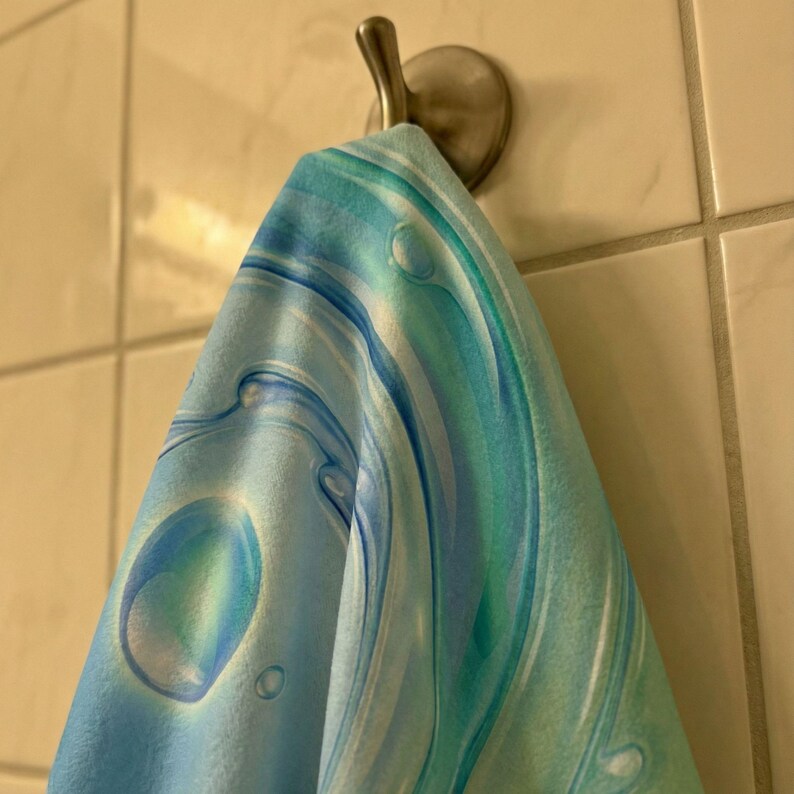 May include: A blue and teal patterned towel hangs from a silver hook on a tiled wall. The towel features a swirling design with bubble-like shapes, creating a water-like effect. The color palette includes shades of blue and green.
